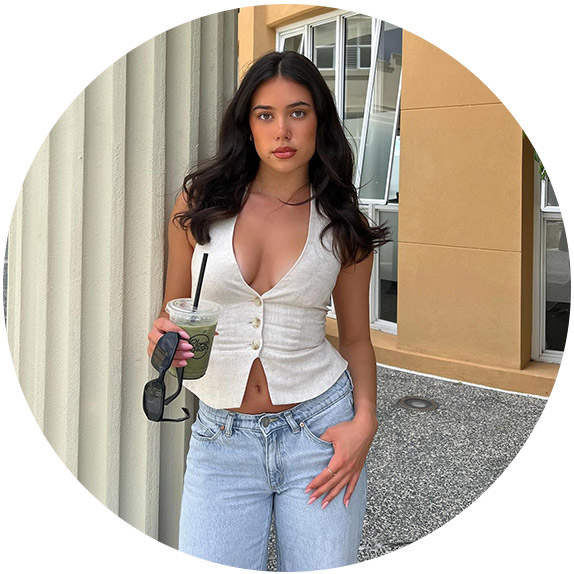 Woman wearing a white halter vest and jeans. She is holding an iced coffee and sunglasses.