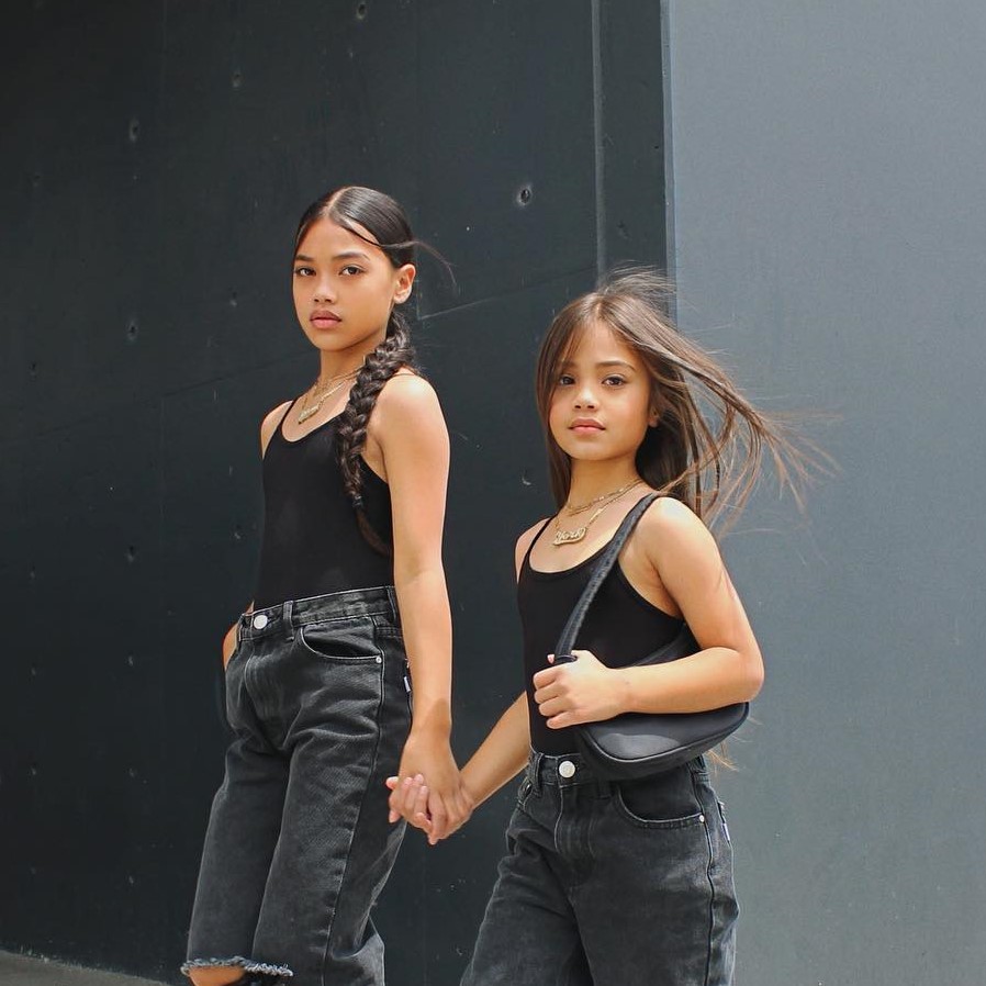 Two young girls wearing matching black tops and jeans, holding hands and posing for the camera