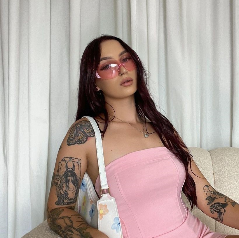 Brunette woman wearing a pink stapless top. She has tattoos on her arms.